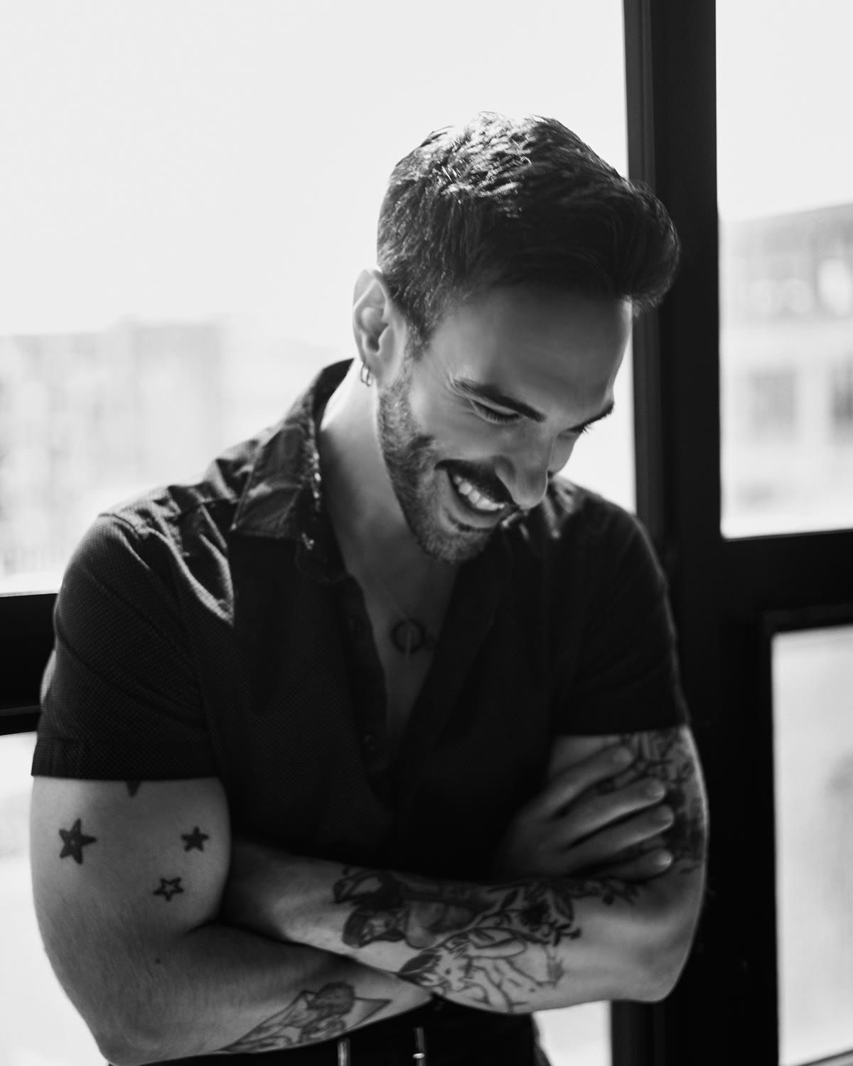 Peter Andrew Danzig in black and white, arms crossed and laughing, wearing a short sleeved shirt with star tattoos visible on one arm, beside a tall window.