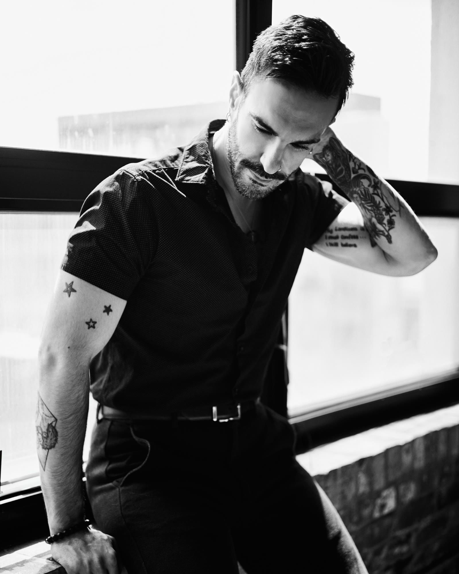 Peter Andrew Danzig in black and white, leaning against a window ledge in a short sleeved shirt, head lowered, star and sleeve tattoos visible on both arms.