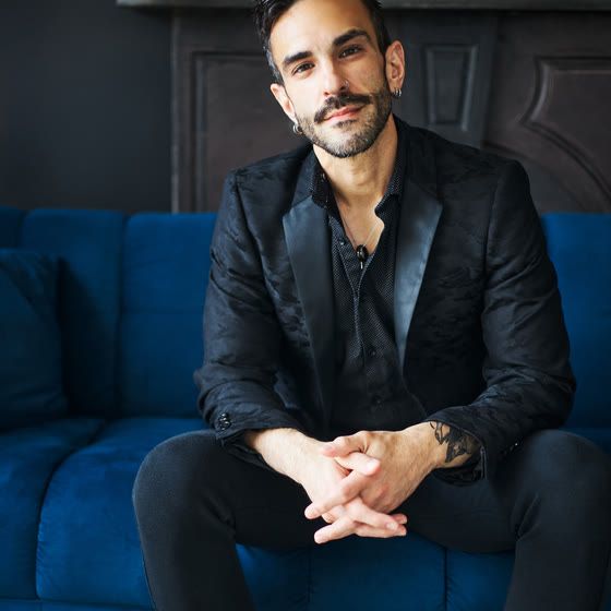 Peter Andrew Danzig seated on a blue velvet sofa in a black jacket, looking directly at the camera.