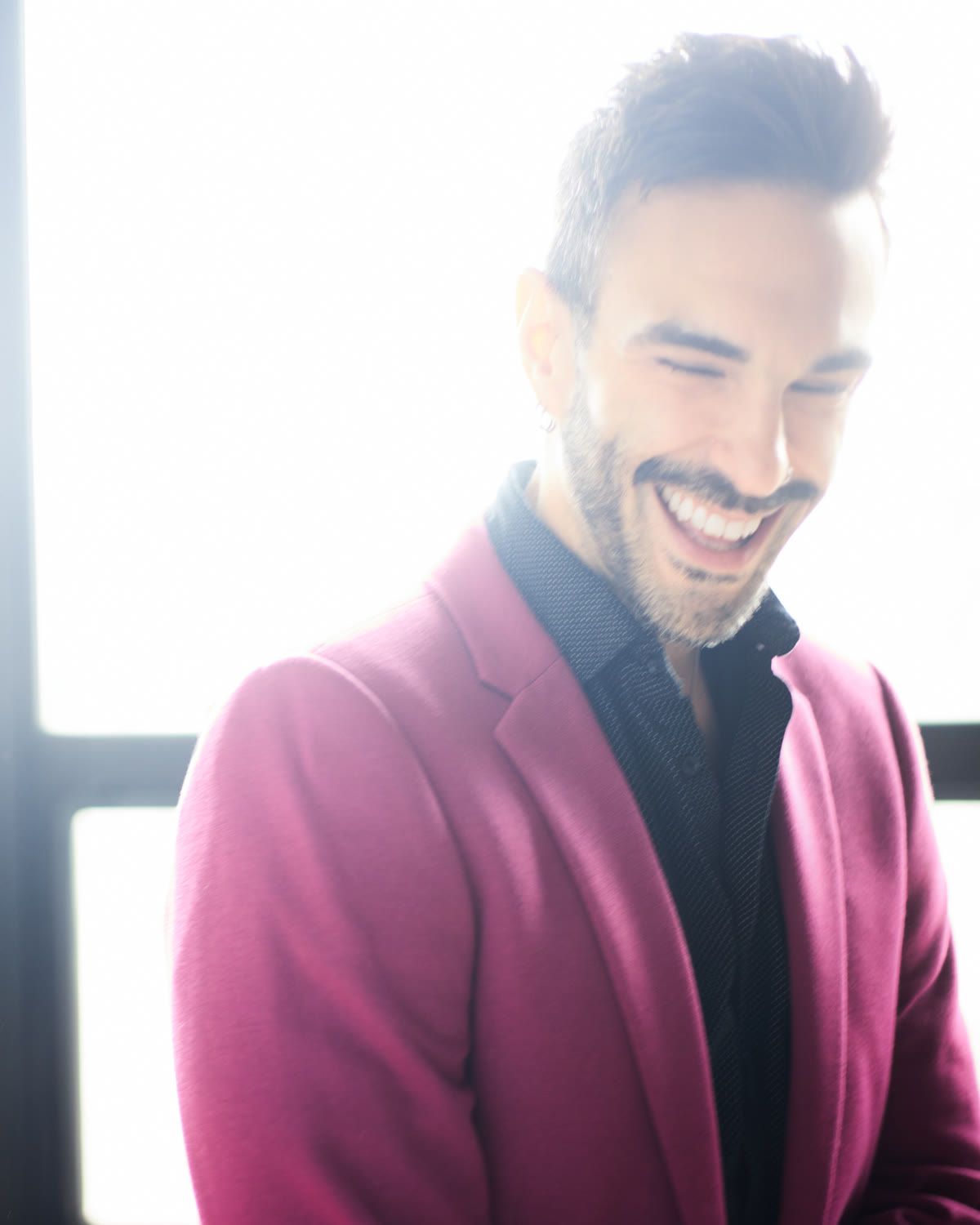 Peter Andrew Danzig in a fuchsia blazer over a dark shirt, head lowered and caught mid laugh, in front of a bright window.
