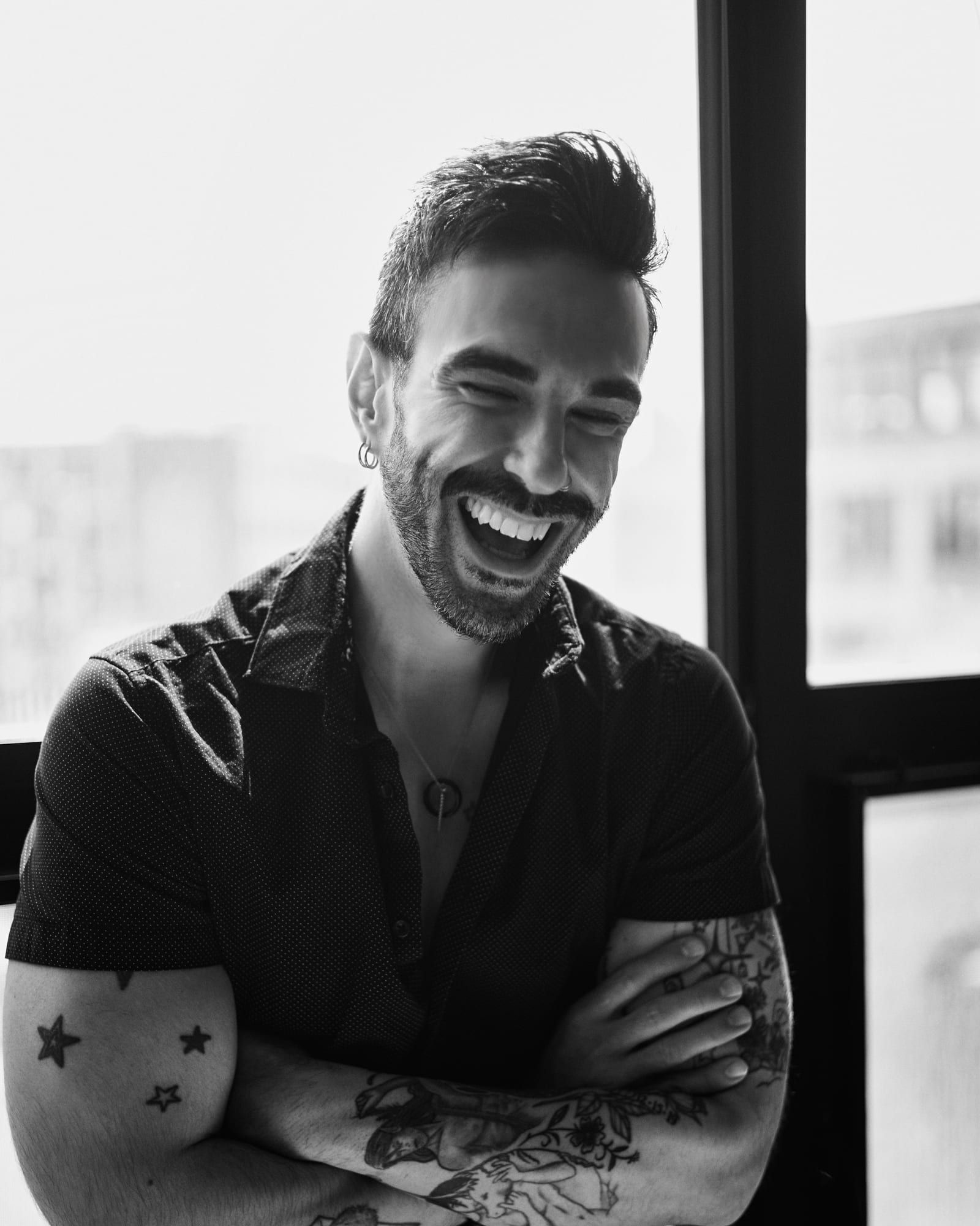 Peter Andrew Danzig in black and white, arms crossed and laughing with his head tilted back, beside a tall window.