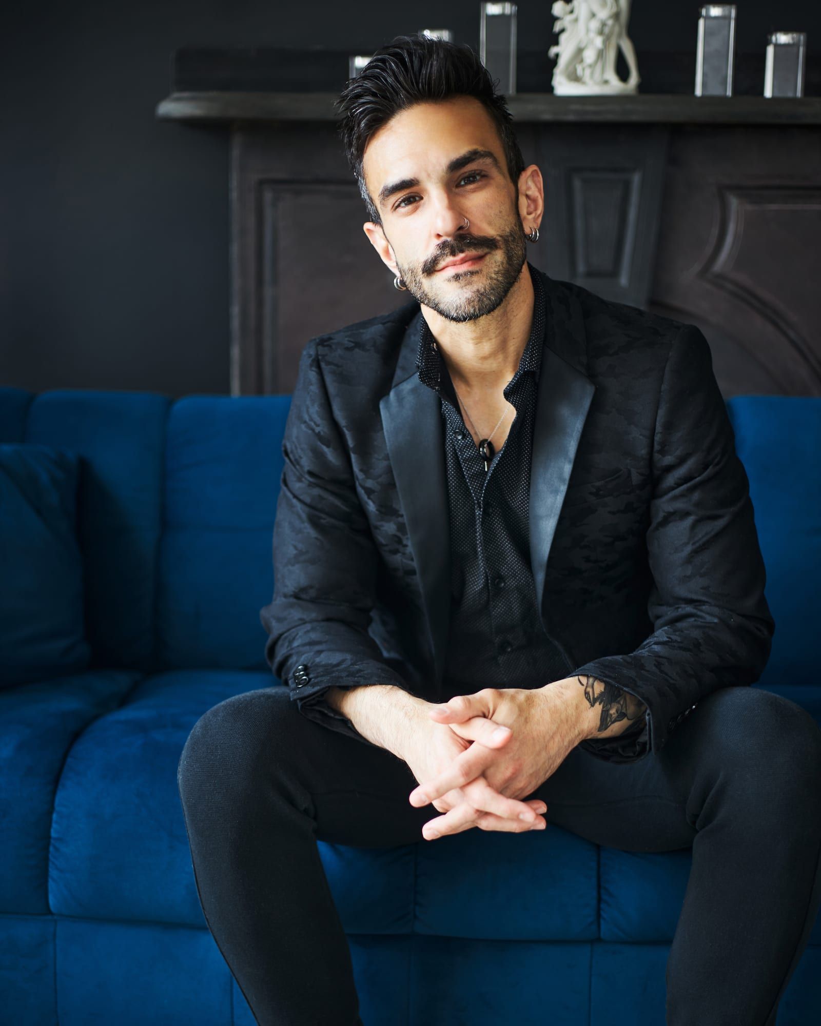 Peter Andrew Danzig seated on a blue velvet sofa in a black jacket and black shirt, hands clasped, looking directly at the camera with a composed expression.