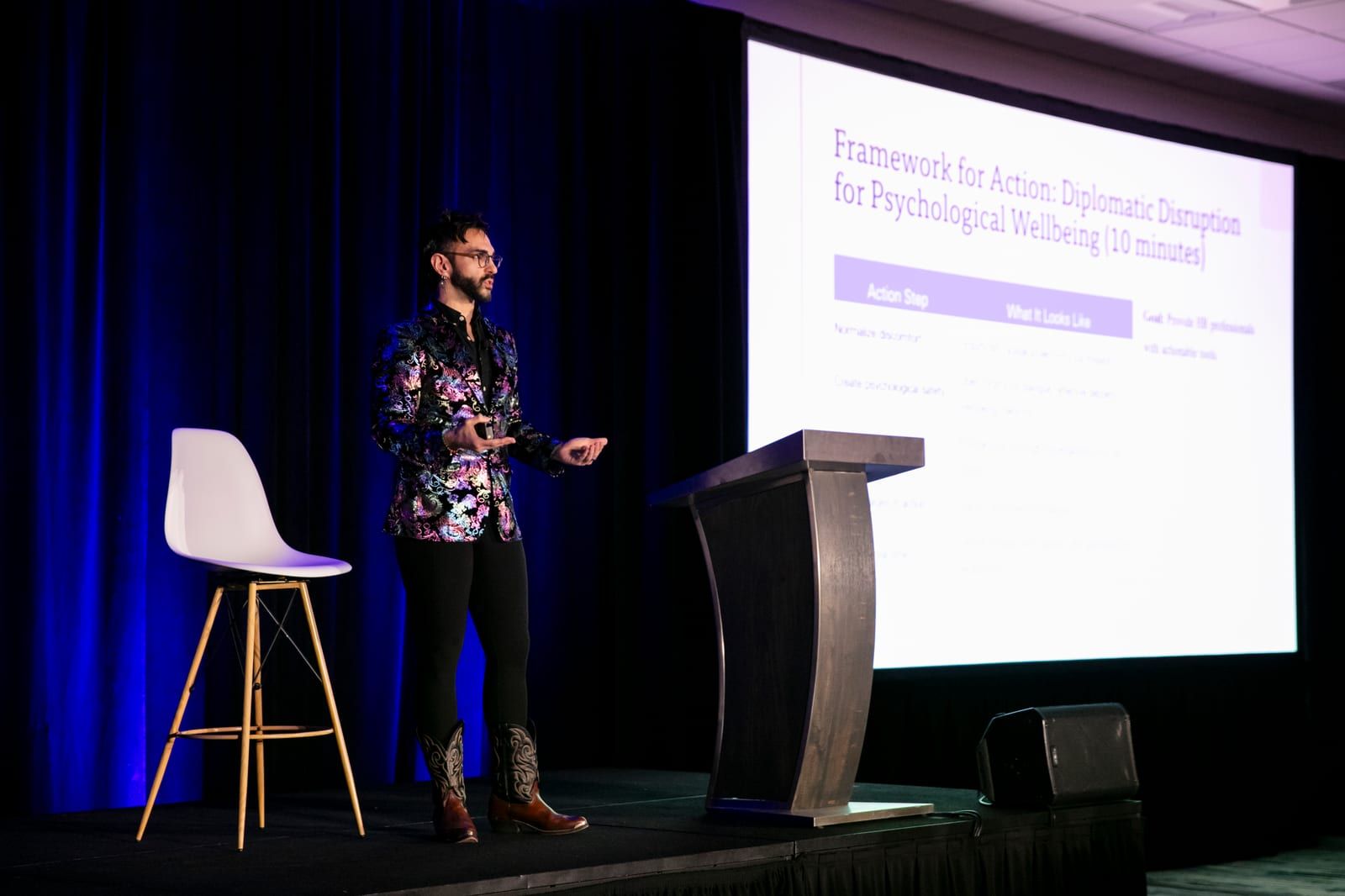 Peter Andrew Danzig speaking on a conference stage in an iridescent floral blazer and cowboy boots, gesturing beside a lectern, with a slide projected behind them reading Framework for Action: Diplomatic Disruption for Psychological Wellbeing.